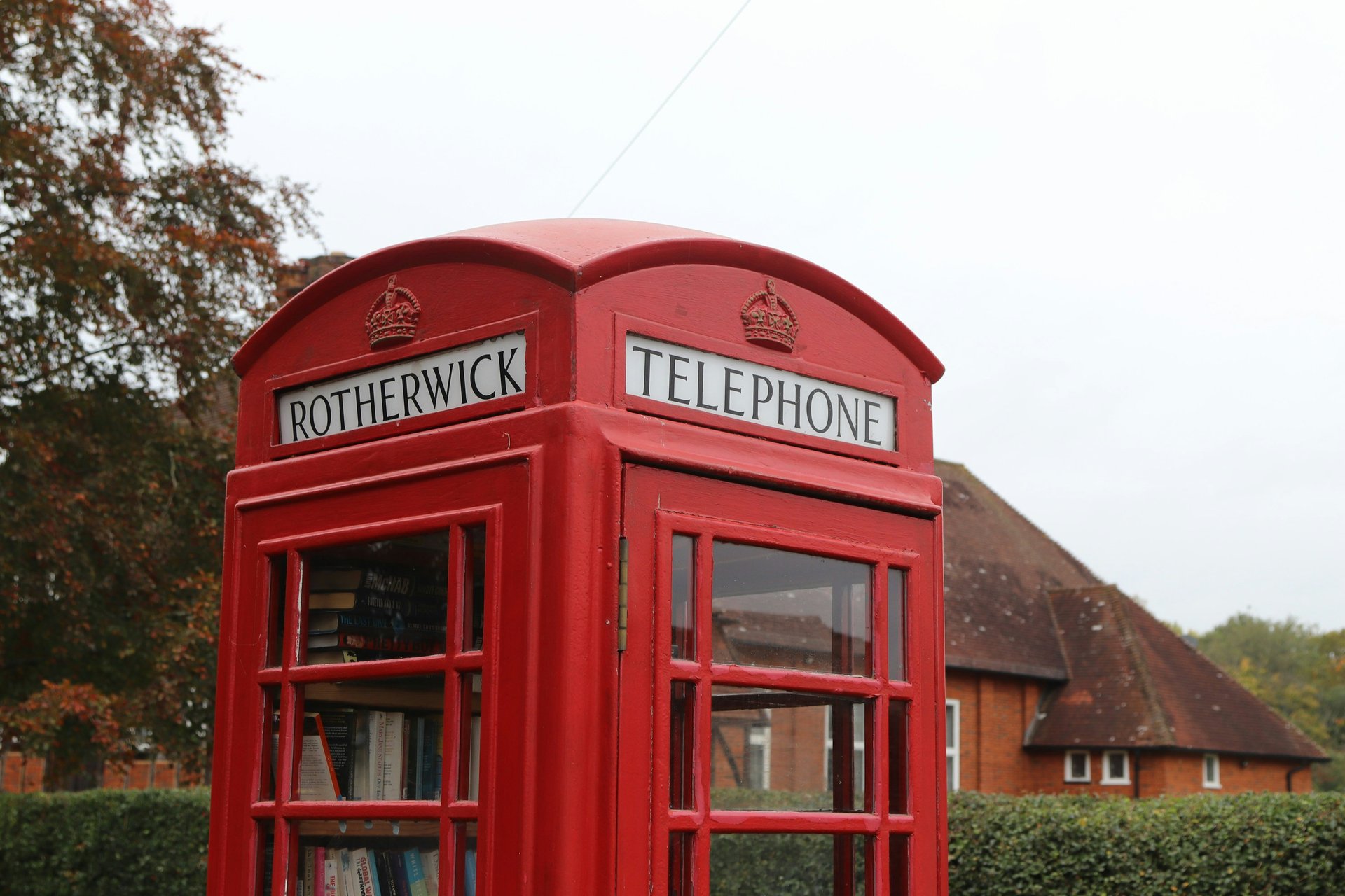 A red British telephone booth with trees and a building behind it